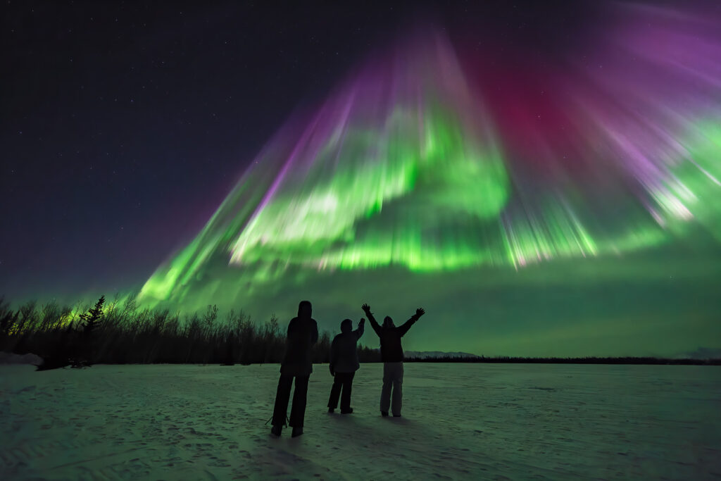 Aurora chasing tour Fairbanks guests witnessing dramatic purple and green corona burst on frozen Alaska lake during intense northern lights geomagnetic storm