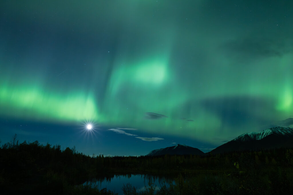 Green aurora borealis sweeping across the Alaska night sky with a starburst moon rising above a still pond, silhouetted boreal forest, and snow-capped mountains reflected in the water.