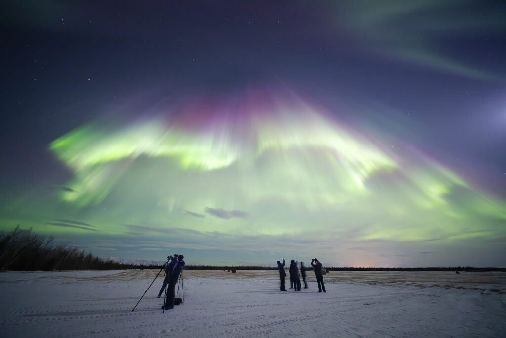 Small group of photographers and aurora watchers silhouetted beneath a dramatic green and magenta aurora corona overhead, standing on snow-covered terrain near Fairbanks, Alaska.