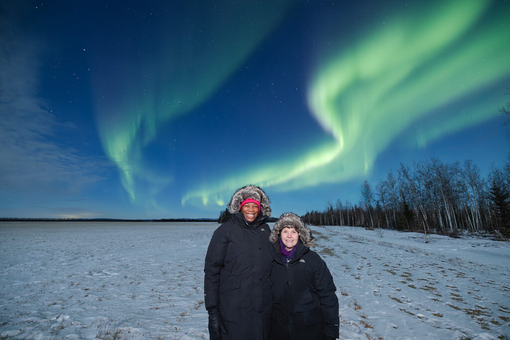 Aurora viewers bundled in extreme cold weather gear photographing northern lights in sub-zero temperatures demonstrating the importance of proper layering and insulated boots