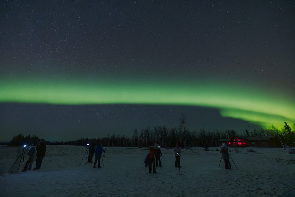 Small group of aurora tour guests with cameras on tripods photographing northern lights while local guide assists with warm tour van visible nearby showing benefits of guided aurora tours