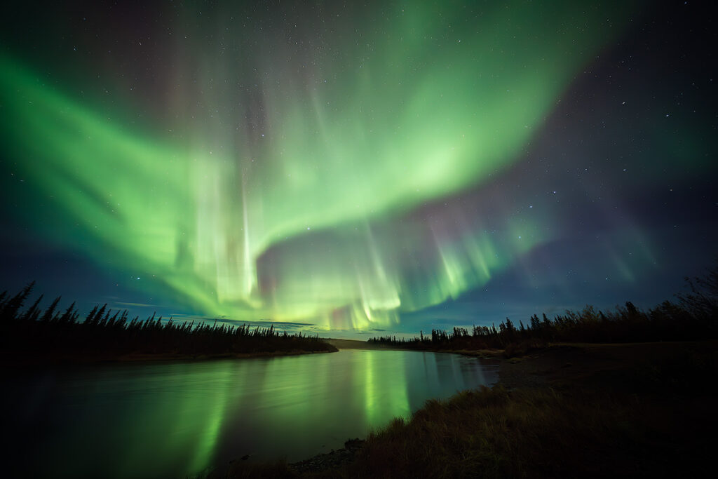 Green northern lights reflecting perfectly on calm lake near Fairbanks in September with fall foliage along shoreline showing ideal conditions for aurora reflection photography