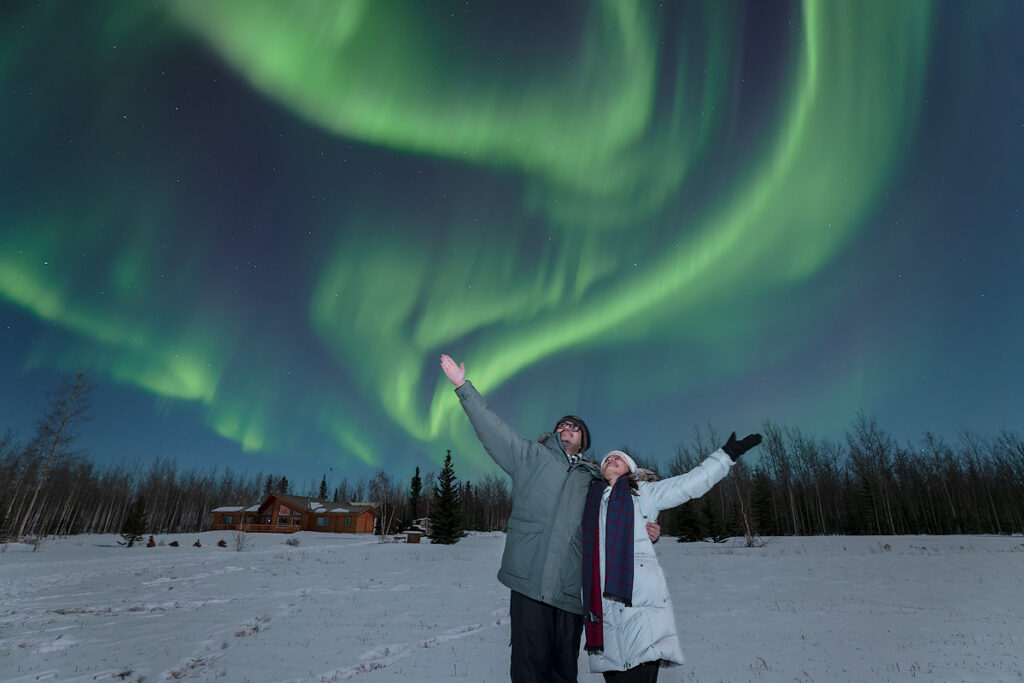 Small group of guests watching vibrant green and purple northern lights from Face The Outdoors private aurora viewing lodge in Fairbanks Alaska.