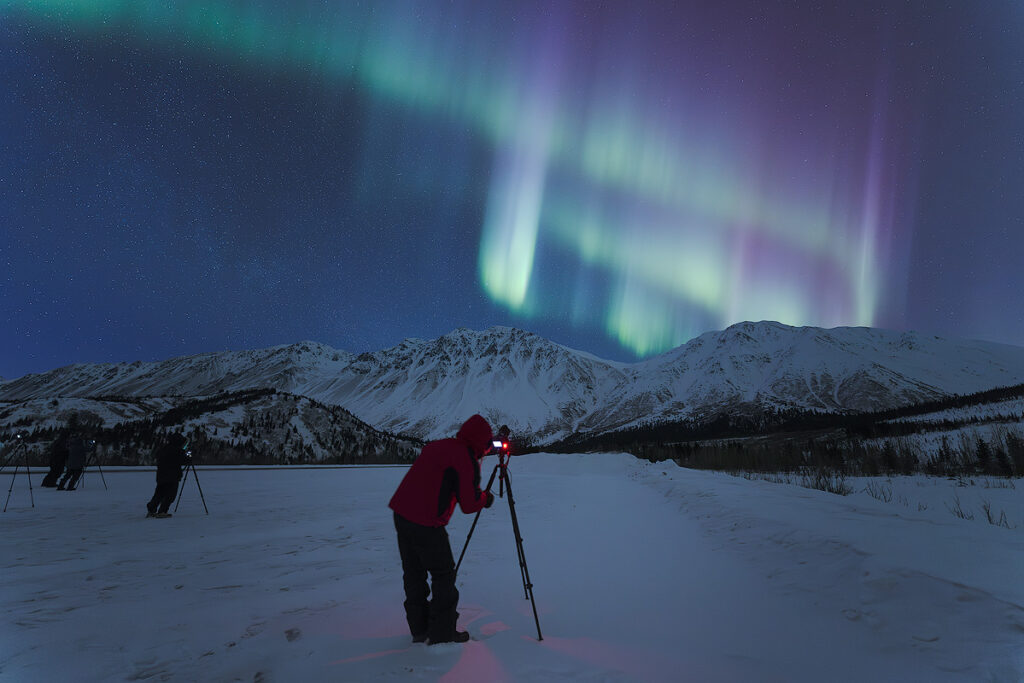 Aurora photography settings — camera on tripod capturing vivid northern lights in Fairbanks using long exposure to reveal human eye aurora colours invisible in real time